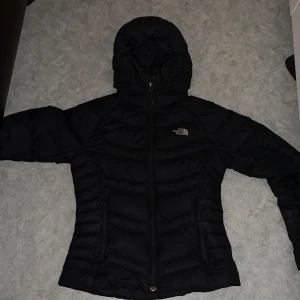 Svart dunjacka från The North Face - Säljer en svart dunjacka från The North Face i mycket bra skick. Jackan har en huva och dragkedja framtill, perfekt för kyliga dagar. Den är stilren och passar både till vardags och utomhusaktiviteter. Storleken är inte specificerad, men den ser ut att ha en normal passform.