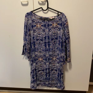 Blå mönstrad klänning från Indiska - Säljer en superfin blå klänning från Indiska i storlek S. Den har ett detaljerat mönster i vitt och orange och är perfekt för både vardag och fest. Klänningen har trekvartsärmar och en knappdetalj i nacken. Materialet känns lätt och bekvämt. Passar perfekt till vår och sommar!