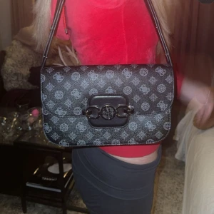 Svart mönstrad axelväska, GUESS - Snygg svart axelväska med grått mönster och en cool spänndetalj på framsidan. Väskan har en praktisk dragkedja på baksidan och justerbar axelrem. Perfekt för både vardag och fest! Från märket GUESS.