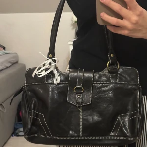 Svart axelväska i skinn - Säljer en snygg svart axelväska med vita sömmar och en dekorativ rem med spänne framtill. Väskan har två rejäla handtag och en praktisk dragkedja upptill. Perfekt för både vardag och fest! 🖤