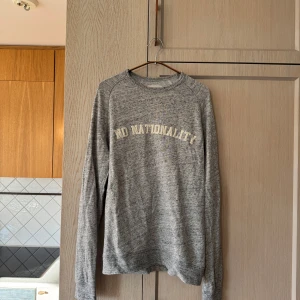 Grå sweatshirt från NN07 - Säljer en snygg grå sweatshirt från NN07 med texten 'No Nationality' tryckt på framsidan. Tröjan är i storlek S och är tillverkad av 60% bomull och 40% polyester, vilket gör den både bekväm och hållbar. Perfekt för en avslappnad stil!