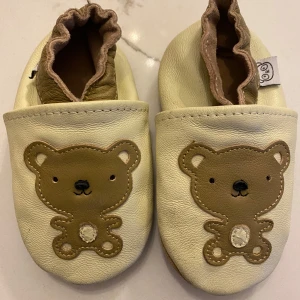 Söta babyskor med björnmotiv - Supergulliga babyskor i beige skinn med ett sött björnansikte på framsidan. Perfekta för små fötter och har en mjuk sula för bekvämlighet. Skorna är i mycket bra skick och passar perfekt för inomhuslek. 12-18 månader.