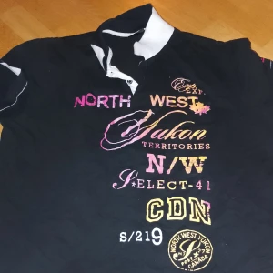 Svart t-shirt med tryck - Säljer en svart t-shirt med coolt tryck i rosa, gult och vitt. Den har en klassisk krage och korta ärmar. Perfekt för en avslappnad stil eller en utekväll. Storleken är inte angiven men verkar vara en herrstorlek.