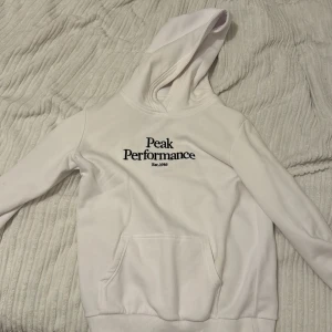 Vit hoodie från Peak Performance - Säljer en stilren vit hoodie från Peak Performance. Den har en klassisk design med loggan broderad på framsidan. Perfekt för både vardag och träning. Hoodien har en stor ficka fram och är i mycket bra skick. Passar perfekt för höst och vår! 😍🤍