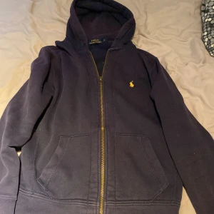 Mörkblå hoodie från Polo Ralph Lauren - Säljer en snygg mörkblå hoodie från Polo Ralph Lauren i storlek S. Den har en dragkedja framtill och en liten broderad logga på bröstet. Tillverkad i mjuk bomull, perfekt för höst och vår. Den är i mycket bra skick och redo för ny ägare
