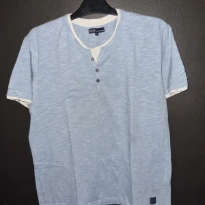 Ljusblå t-shirt från S.C.W. - Säljer en ljusblå t-shirt från S.C.W. i storlek L. Den har en avslappnad stil med korta ärmar och en knappdetalj vid halsen. 