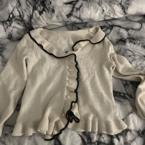 Vit kofta med pärldetaljer - Superfin vit kofta med svarta kanter och pärlknappar. Den har en söt volangdetalj längs knäppningen och är perfekt för både vardag och fest. Koftan är i ett mjukt material och har långa ärmar. Passar perfekt till höst och vår!