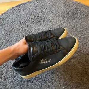 Säljer ett par snygga svarta sneakers från Saint Laurent i storlek 45 och mycket bra skick. De har en stilren design med svart skinn och en kontrasterande beige sula. Perfekta för både vardag och fest! Hör av er om ni e intresserade