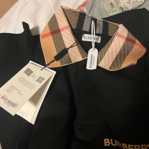 Svart pikétröja från Burberry - Säljer en snygg svart pikétröja från Burberry med deras ikoniska rutiga mönster på kragen. Tröjan är i nytt skick och har en klassisk passform. Perfekt för både vardag och fest! 🖤