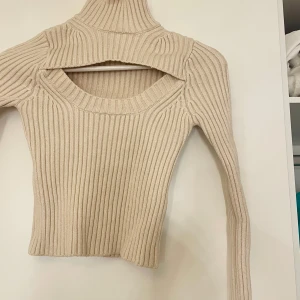 Beige ribbad tröja från Bershka - Säljer en superfin beige ribbad tröja från Bershka. Den är croppad och har en unik design med en öppen detalj vid halsen. Perfekt för höst och vår, och den är både stilren och bekväm. Passar perfekt till både jeans och kjol!