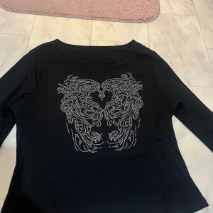 Svart långärmad topp med strassdetaljer - Snygg svart långärmad topp med glittrande strassdetaljer i form av ett mönster framtill. Toppen har en v-ringad hals och är perfekt för att ge en extra touch till din outfit.