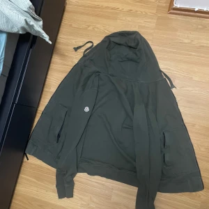 Grön hoodie från Moncler - Säljer en snygg grön hoodie från Moncler med huva och dragsko. Den har en broderad Moncler-logga på bröstet och är perfekt för en avslappnad stil. Hoodien har långa ärmar och en bekväm passform.