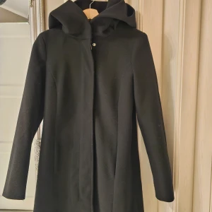 Svart kappa med stor huva från Vero Moda - Snygg svart kappa från Vero Moda med stor huva. Tecken på användning finns, dvs noppror på armar och lite på sidorna, kanske lätt att ta bort med noppborttagare? Utöver det, väldigt fin & skön!