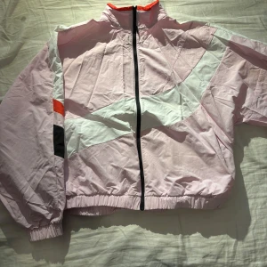 Rosa vindjacka från Reebok - Säljer en rosa vindjacka från Reebok med dragkedja och färgblock i vitt, svart och röd. Jackan har långa ärmar och elastiska muddar. Perfekt för utomhusträning eller bara vardagligt. Den är lite kortare i längden därav har jag köpt storlek L.