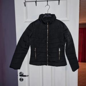 Svart pufferjacka med dragkedja - Säljer en stilren svart pufferjacka med dragkedja framtill och två praktiska fickor med dragkedjor. Jackan har en hög krage och är perfekt för kyligare dagar. Den quiltade designen ger en modern look. Nyskick,  endast prövad.