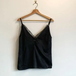 Svart linne från Vera & Lucy - Snygg svart linne från Vera & Lucy i 100% polyester med spetsdetaljer längs v-ringningen. Perfekt för en stilren look. Justerbara axelband för bästa passform.