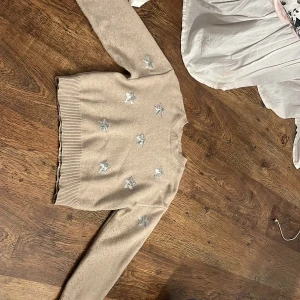 Beige stickad tröja från Gina Tricot - Säljer en söt beige stickad tröja från Gina Tricot med silverfärgade paljettstjärnor på framsidan. Tröjan har långa ärmar och ribbade muddar vid ärmslut och nederkant. Perfekt för att lysa upp vardagen! ✨
