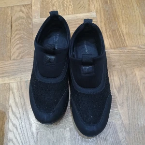 Svarta sneakers med glittrig detalj - Snygga svarta sneakers med glittrig ovandel och elastisk passform. Perfekta för en stilren look med en touch av glitter. Skorna har en bekväm sula och är lätta att ta på och av.