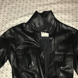 Svart skinnjacka från Zara - Snygg svart skinnjacka från Zara med knappar och bälte i midjan. Jackan har långa ärmar och två bröstfickor med lock. Perfekt för en stilren look.