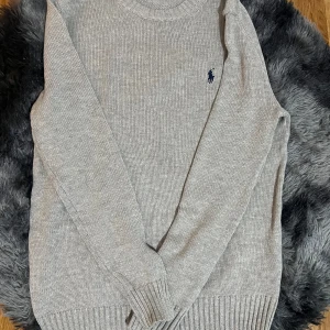 Ralph Lauren tröja  - Säljer en stilren grå stickad tröja från Polo Ralph Lauren med det klassiska logotypbroderiet på bröstet. Tröjan har långa ärmar och ribbade muddar vid ärmslut och nederkant. Perfekt för kyligare dagar!