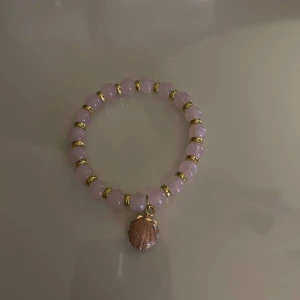 Rosa armband med snäckberlock - Charmigt rosa armband med glittriga pärlor och en snäckformad berlock i guld. Perfekt för att ge en somrig touch till din stil.