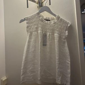 Vit ärmlös smockad topp från Fiore!!! - Säljer en söt vit ärmlös topp från Fiore med smockdetaljer vid halsen och axlarna. Tillverkad i 100% linne och därmed priset. Original priset är 1100! Kan möjligtvis diskutera pris💕Strlk M men funkar för både S och M. Aldrig använd