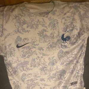 Vit och blå Frankrike tröja - Snygg vit fotbollströja från Nike med ett unikt blått mönster och en tupp på bröstet. Tröjan har korta ärmar och är perfekt för fotbollsfans som vill sticka ut. Den har en modern design med en sportig känsla.