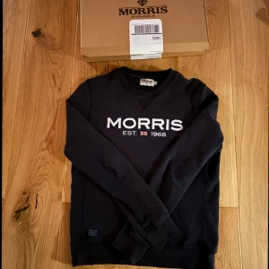 Svart tröja från Morris - Säljer en stilren svart tröja från Morris med texten 'Morris EST. 1968' tryckt på framsidan. Tröjan har långa ärmar och en rund halsringning. Perfekt för en avslappnad och trendig look.