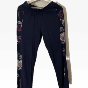 Svarta byxor med blommönster - Snygga svarta byxor med ett färgglatt blommönster längs sidorna. Perfekta för att lägga till en touch av stil till din garderob. Byxorna har en bekväm passform och är gjorda i ett mjukt material. Ej använd 