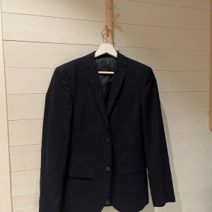 Mörkblå kavaj - Elegant mörkblå kavaj med två knappar framtill och klassisk krage. Kavajen har långa ärmar med fyra dekorativa knappar vid ärmsluten. Perfekt för en stilren look.