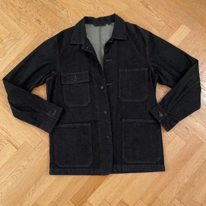 Dark Denim Overshirt - Snygg mörkblå overshirt i jeans med knappar framtill och flera praktiska fickor, bl.a innerficka. OANVÄND. Perfekt för våren och sommarn. Kommer från Uniqlo. Det står S men den passar som M. Den är tillverkad i slitstarkt denimtyg. 