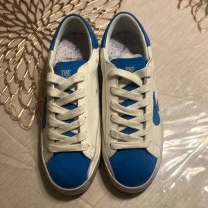 Vita och blå sneakers från The Gallerist - Snygga vita sneakers med blå detaljer från The Gallerist. Skorna har en unik design med blå klubbsymboler på sidorna och text på hälen. Perfekta för en stilren look. Storlek 37. Dem är aldrig använda och kan mötas upp vid antingen Kungsbacka eller Mölndal ifall ni inte vill ta frakt. 