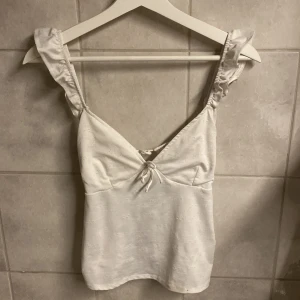 Vit topp med volangdetaljer från Divided - Säljer en söt vit topp från Divided med volangdetaljer på axelbanden och en knytning framtill. Perfekt för varma dagar och kan enkelt matchas med olika outfits. Toppen är ärmlös och har en feminin design.