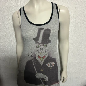 Grå ärmlös linnetopp med tryck - Snygg grå ärmlös linnetopp med ett unikt tryck av en skelettfigur i kostym och hög hatt. Toppen har svarta kanter vid ärmhål och halsringning. Perfekt för en cool och avslappnad stil.