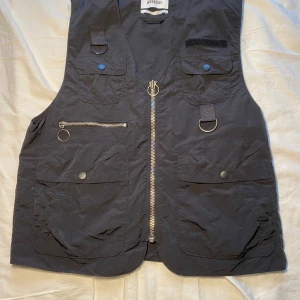 Svart väst från Weekday - Snygg svart väst från Weekday med flera praktiska fickor och dragkedja framtill. Perfekt för en cool och avslappnad stil. Västen har även dekorativa ringdetaljer och en dragkedja på baksidan. Knapparna är inte blåa, ljuset får de att se ut så.