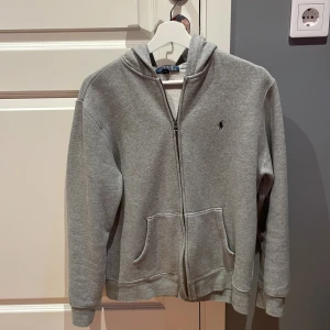 Grå hoodie från Polo Ralph Lauren - Säljer en stilren grå hoodie från Polo Ralph Lauren med dragkedja och broderad logga på bröstet. Tröjan har krymp i tvätten så passar folk som har storlek xs/s