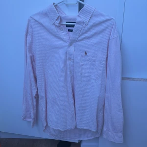 Rosa randig skjorta från Polo Ralph Lauren - Säljer en stilren rosa och vit randig skjorta från Polo Ralph Lauren. Skjortan är i 100% bomull och har långa ärmar samt en klassisk krage. Den har en broderad logotyp på bröstfickan och knappar framtill. Perfekt för en avslappnad men ändå elegant look. Det är en gammal model av ralph Lauren skjortor och ganska unik 