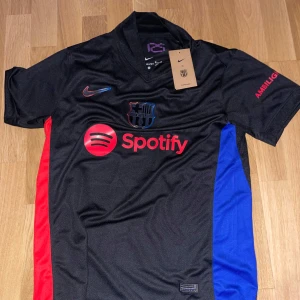 Svart FC Barcelona fotbollströja från Nike - Snygg svart FC Barcelona tröja från Nike med korta ärmar. Tröjan har röda och blå detaljer på sidorna och klubbens emblem på bröstet. Den är märkt med Spotify och UNHCR-loggor. Perfekt för fans som vill visa sitt stöd. Mycket snygg tröja. Helt ny ej använd! Affär kan diskuteras vid snabb affär!