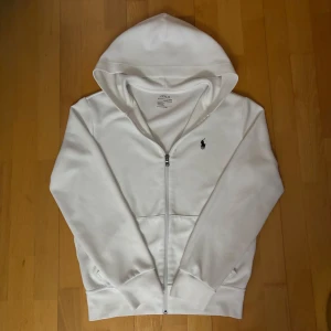 Vit hoodie från Ralph Lauren - Säljer en vit hoodie från Ralph Lauren med dragkedja och klassisk logga på bröstet. Perfekt för en avslappnad look. Den har en bekväm passform och är tillverkad i mjukt material. Det är storlek XS men passar även S. Snören saknas så Priset är förhandlingsbart!