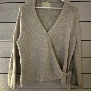 Grå omlottkofta - Säljer en mysig grå omlottkofta med knytning i sidan. Koftan har långa ärmar och en v-ringad design. 