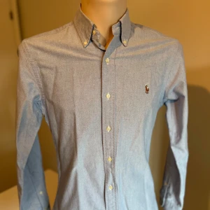 Blå skjorta från Ralph Lauren - Snygg blå skjorta från Ralph Lauren med klassisk krage och broderad logga på bröstet. Skjortan har långa ärmar och knappar fram samt vid manschetterna. Perfekt för en stilren look. Mycket bra skick. Nypris 1600-1900kr