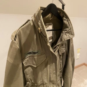 Field Jacket från Ralph Lauren - Säljer en grön jacka från Ralph Lauren's Denim & Supply. Jackan har flera fickor och en dragkedja framtill. Den har en cool militärinspirerad look med broderade detaljer på bröstet. Perfekt för en avslappnad stil.