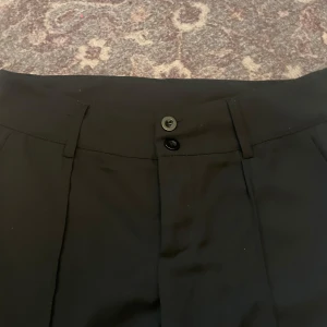 Svarta kostymbyxor - Snygga svarta kostymbyxor med hög midja och raka ben. Byxorna har en klassisk design med knappar framtill och är perfekta för en stilren look. 