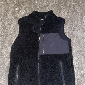 Svart fleeceväst med dragkedja - Säljer en svart fleeceväst från SMOG med dragkedja och en praktisk bröstficka. Västen har en stilren design och är perfekt för lager-på-lager under kyligare dagar. Storlek xs med passar även S