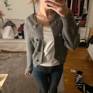 Grå kofta med knappar - Säljer en stilren grå kofta med knappar framtill. Koftan har ett diskret mönster och långa ärmar. Perfekt för att hålla sig varm och snygg under kyligare dagar.
