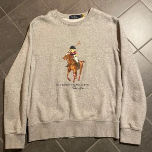 Grå sweatshirt från Ralph Lauren - Säljer en grå sweatshirt från Ralph Lauren med ett stort tryck av en polospelare på framsidan. Tröjan har rund hals och långa ärmar, perfekt för en avslappnad stil. Klassisk och stilren design som passar till mycket. Qr koden på tröjan leder till hemsidan som du ser på femte bilden. 