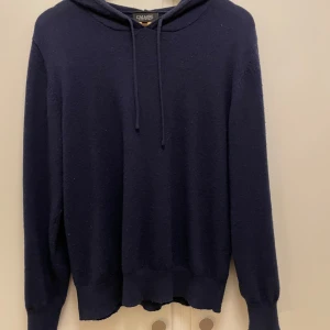 Mörkblå cashmere hoodie från Calmire - Säljer en lyxig och mjuk mörkblå hoodie i cashmere från Calmire. Perfekt för kyliga dagar med sin sköna huva och långa ärmar. En stilren och bekväm tröja för alla tillfällen. Storlek M men passar S