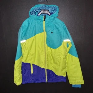 Flerfärgad jacka från H&M - Säljer en färgglad jacka från H&M i blått, gult och lila. Jackan har en praktisk huva och dragkedja framtill. Perfekt för kyliga dagar med sin långa ärmar och reflekterande detaljer för extra synlighet. Har ett litet hål på ena axeln,  tror att den händiga fixar de lätt.