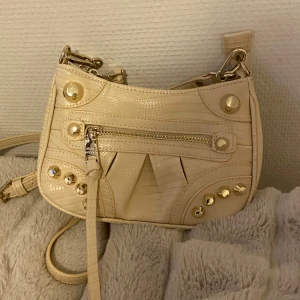 Beige axelväska med nitar - Snygg beige axelväska med coola nitar och dragkedja framtill. Väskan har en justerbar axelrem och är perfekt för att ge din outfit en edgy touch. Väskan har aldrig kommit till användning så därför säljs den