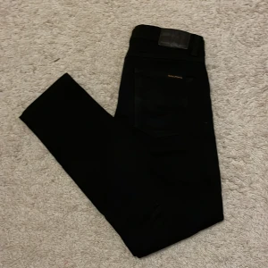 Svarta jeans från Nudie Jeans - Snygga svarta jeans från Nudie Jeans med klassisk femficksdesign. De har en diskret broderad detalj på bakfickan och en läderpatch med logotyp vid midjan. Perfekta för en stilren look.